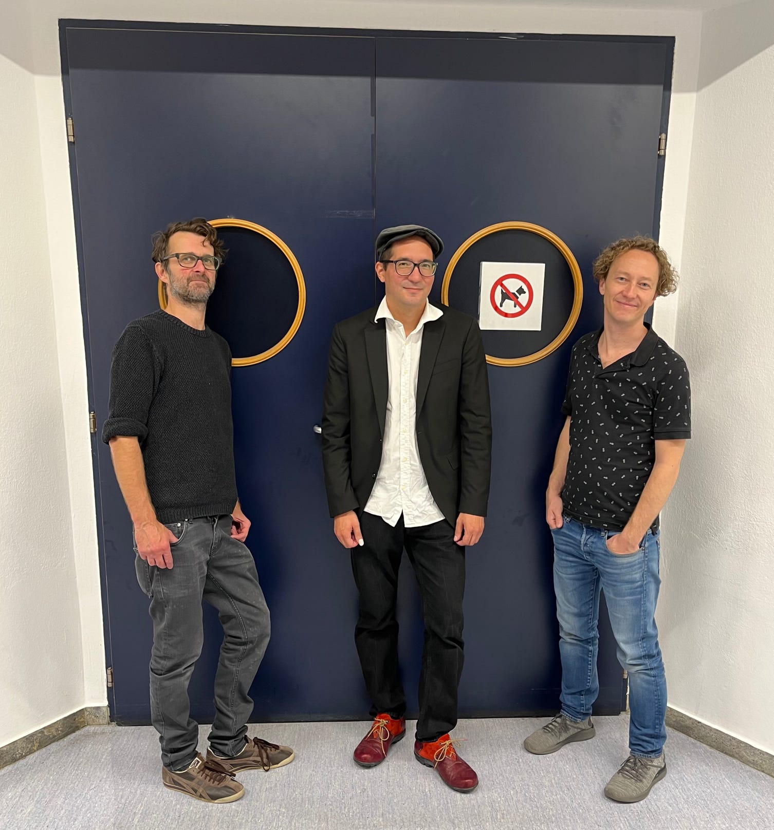 Die Band MRT. Von Links nach Rechts: Stefan Thaler, Michiru Ripplinger und Thomas Froschauer. Die Musiker stehen vor einer Doppeltür im Theater am Steg in Baden, eine Stadt im Süden von Wien. Ein Hundeverbotschild ist auch zu entdecken.